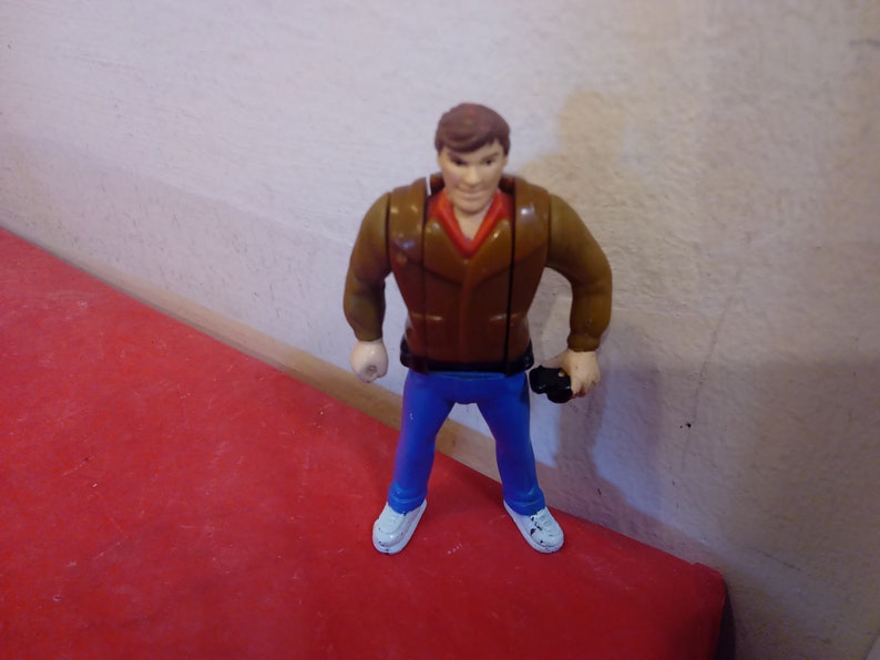 Pode incluir: Uma jaqueta de couro marrom com uma camisa vermelha por baixo, jeans azul e t&ecirc;nis branco. A figura est&aacute; segurando um objeto preto na m&atilde;o direita.