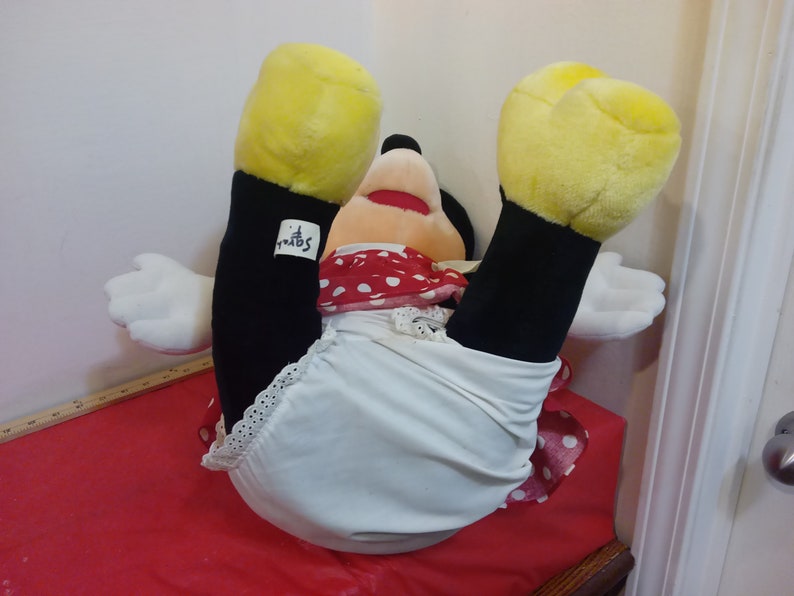 Puede incluir: Un juguete de peluche de Minnie Mouse con un vestido de lunares rojos y blancos y ropa interior blanca. El juguete tiene manos y pies amarillos y ojos negros. El juguete est&aacute; acostado de espaldas con las piernas en el aire. El juguete tiene una etiqueta "Disney" en su pierna.