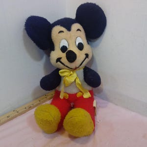 May include: Vintage Mickey Mouse plush toy. The plush features a black head and ears, a cream-colored face, and a yellow bow tie. Mickey wears red pants and yellow shoes. A classic Disney collectible.