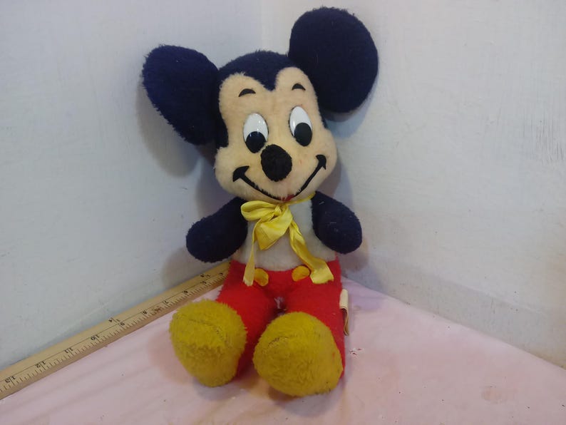 May include: Vintage Mickey Mouse plush toy. The plush features a black head and ears, a cream-colored face, and a yellow bow tie. Mickey wears red pants and yellow shoes. A classic Disney collectible.