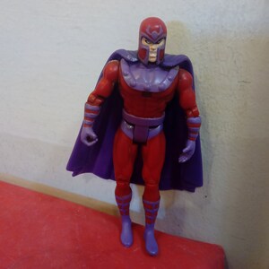 Pode incluir: Uma figura de a&ccedil;&atilde;o vermelha e roxa do Magneto, um super-her&oacute;i da Marvel Comics. A figura tem uma capa roxa e um capacete vermelho com uma viseira roxa.
