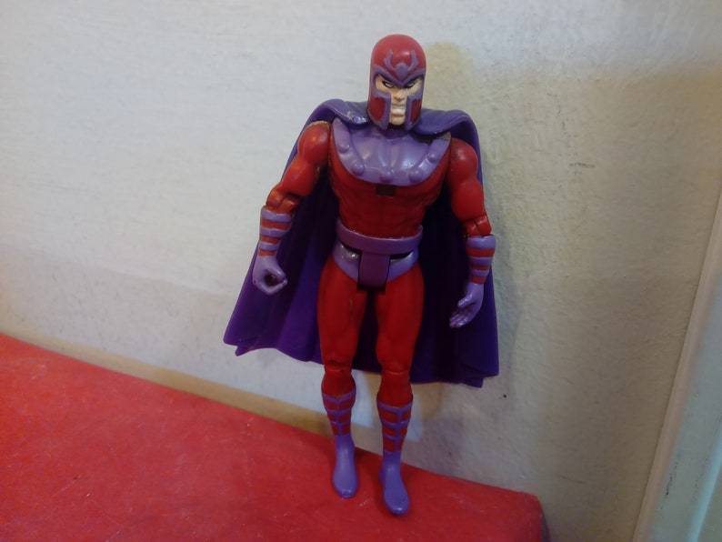 Pode incluir: Uma figura de a&ccedil;&atilde;o vermelha e roxa do Magneto, um super-her&oacute;i da Marvel Comics. A figura tem uma capa roxa e um capacete vermelho com uma viseira roxa.