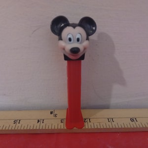 Peut inclure: Une tête de Mickey Mouse en plastique rouge et noir sur un distributeur de bonbons Pez rouge et blanc.