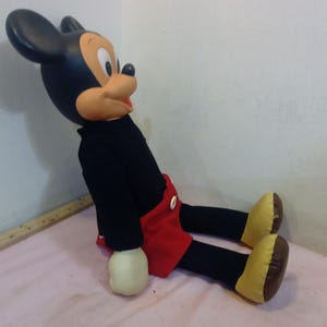 May include: A vintage Mickey Mouse plush doll, featuring a black body, red shorts, and yellow shoes. The doll has a large head with black ears, a tan face, and white gloves. The plush is sitting on a pink surface.