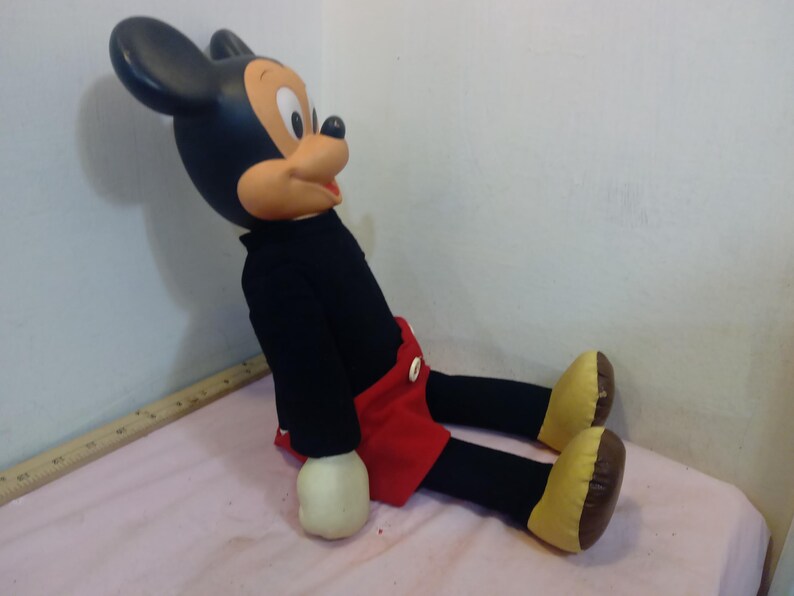 May include: A vintage Mickey Mouse plush doll, featuring a black body, red shorts, and yellow shoes. The doll has a large head with black ears, a tan face, and white gloves. The plush is sitting on a pink surface.
