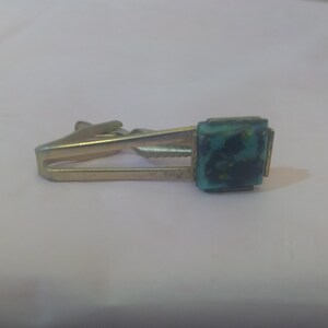 May include: A gold tie clip with a square turquoise blue stone with black and gold flecks.