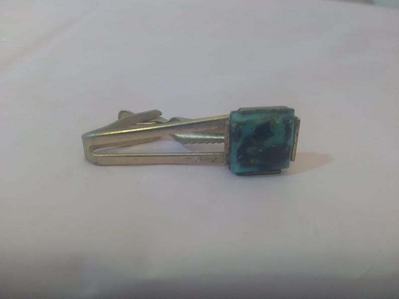 May include: A gold tie clip with a square turquoise blue stone with black and gold flecks.