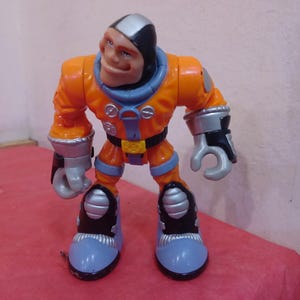 May include: Orange and gray astronaut action figure with a black helmet and a friendly face. The figure has a silver belt and silver details on the boots.