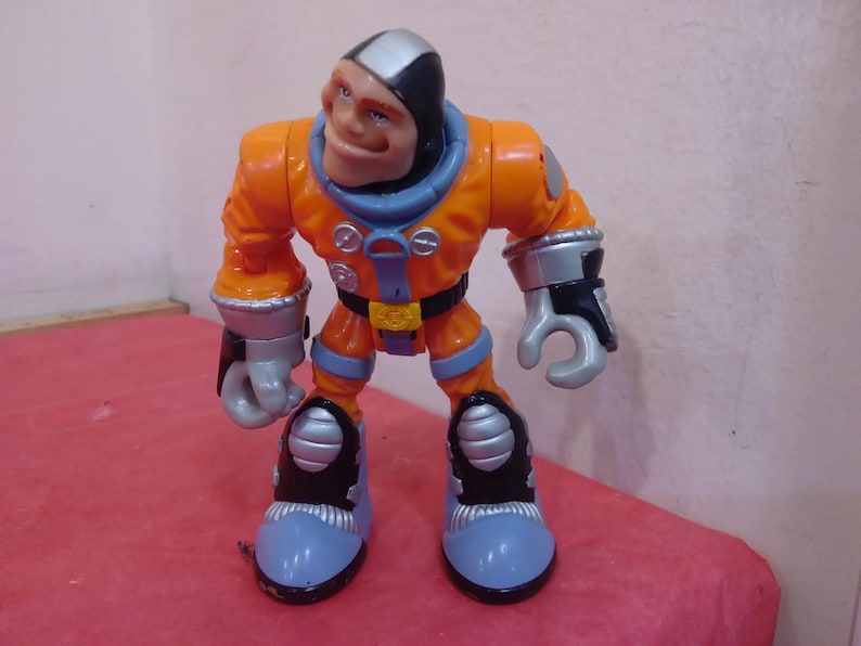 May include: Orange and gray astronaut action figure with a black helmet and a friendly face. The figure has a silver belt and silver details on the boots.