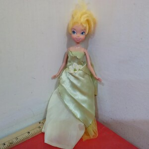 May include: A doll with blonde hair and blue eyes wearing a light green satin gown with a white flower accent. The doll's dress has a gold trim and a yellow underskirt. The doll is standing on a red surface.