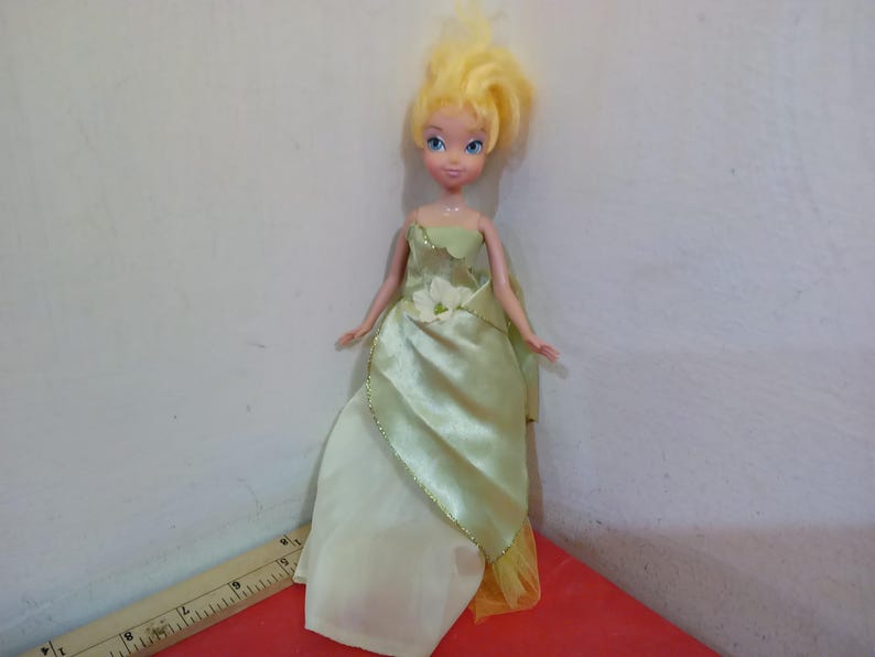 May include: A doll with blonde hair and blue eyes wearing a light green satin gown with a white flower accent. The doll's dress has a gold trim and a yellow underskirt. The doll is standing on a red surface.