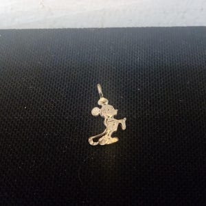 May include: A gold-colored charm featuring a silhouette of Mickey Mouse. The charm is a simple design with Mickey Mouse standing with his arms at his sides.