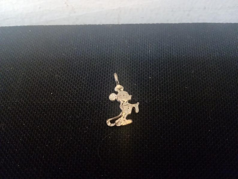 May include: A gold-colored charm featuring a silhouette of Mickey Mouse. The charm is a simple design with Mickey Mouse standing with his arms at his sides.