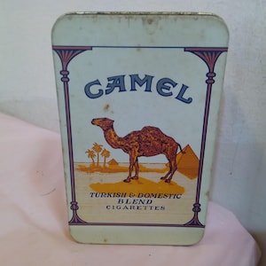 Op de afbeelding: Vintage Camel sigarettenblik met een rechthoekige vorm. Het blik toont een kameelillustratie, palmbomen en piramides. De tekst "CAMEL" staat bovenaan, met daaronder "TURKISH & DOMESTIC BLEND CIGARETTES".