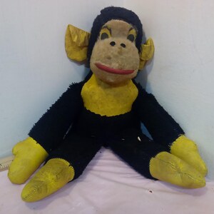 May include: Vintage stuffed monkey toy with black fur, yellow hands, feet, and chest, and a tan face with a red mouth. The monkey has large yellow ears and is sitting down.
