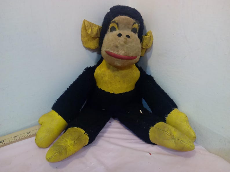 May include: Vintage stuffed monkey toy with black fur, yellow hands, feet, and chest, and a tan face with a red mouth. The monkey has large yellow ears and is sitting down.