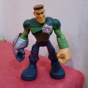 May include: A green and black action figure with a silver arm and a red and yellow symbol on its chest. The figure has a brown head with a sculpted face and short brown hair.