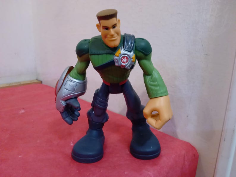 May include: A green and black action figure with a silver arm and a red and yellow symbol on its chest. The figure has a brown head with a sculpted face and short brown hair.