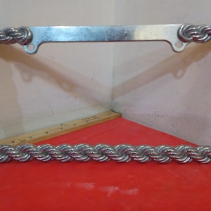 May include: A silver-colored license plate frame with a braided rope design. The frame has a rectangular shape with rounded corners and mounting holes. The braided rope design adds a unique and decorative touch to the frame.
