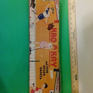 May include: A vintage game box with illustrations of children playing various games. The box is yellow and orange with black text that reads "KRO KAY and FIVE OTHER GAMES" and "ARCH-PIN".