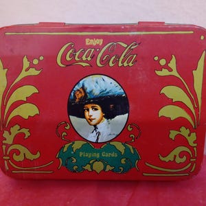 May include: Red metal tin with gold accents and a Coca-Cola logo. The tin features a vintage illustration of a woman wearing a hat and the text "Enjoy Coca-Cola Playing Cards".