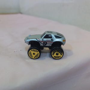 Peut inclure: Petite voiture jouet argentée avec de grandes roues noires et des jantes jaunes. Le numéro 9 et le mot "ERIC" sont visibles sur le côté. Ce jouet est un petit monster truck.