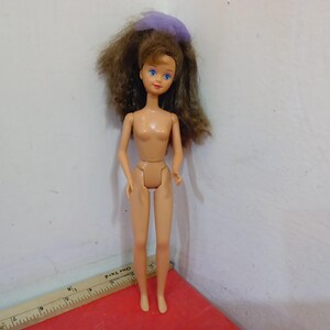 May include: A vintage doll with brown hair and blue eyes. The doll has a light skin tone and is wearing a purple hair accessory. The doll is standing on a red surface.