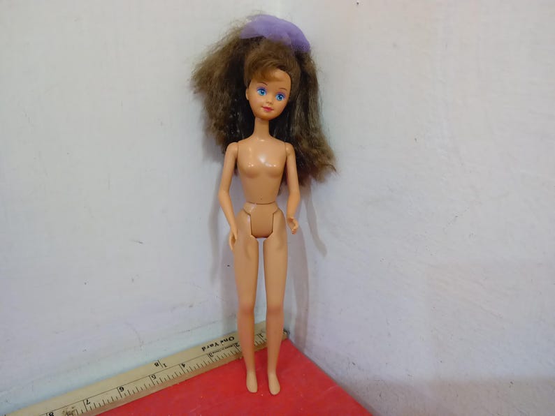 May include: A vintage doll with brown hair and blue eyes. The doll has a light skin tone and is wearing a purple hair accessory. The doll is standing on a red surface.