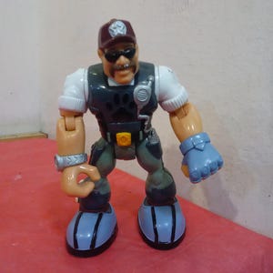 May include: A plastic action figure of a man wearing a brown hat, sunglasses, a white shirt, a dark blue vest, and gray pants. The figure has a paw print on the vest and is holding a walkie-talkie.