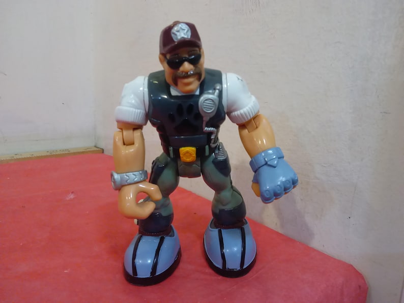 May include: A plastic action figure of a man wearing a brown hat, sunglasses, a white shirt, a dark blue vest, and gray pants. The figure has a paw print on the vest and is holding a walkie-talkie.