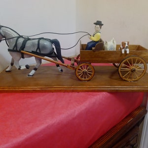 Può includere: Figurina in legno di un carro trainato da cavalli. Il cavallo grigio tira un carro marrone con un conducente, due cani e ruote di legno. La figurina è montata su una base di legno.