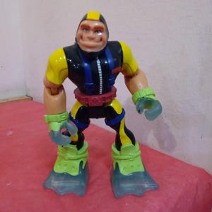 May include: A yellow, black, and blue action figure with a large head and flippers for feet. The figure is wearing a black wetsuit with a zipper and a yellow belt.