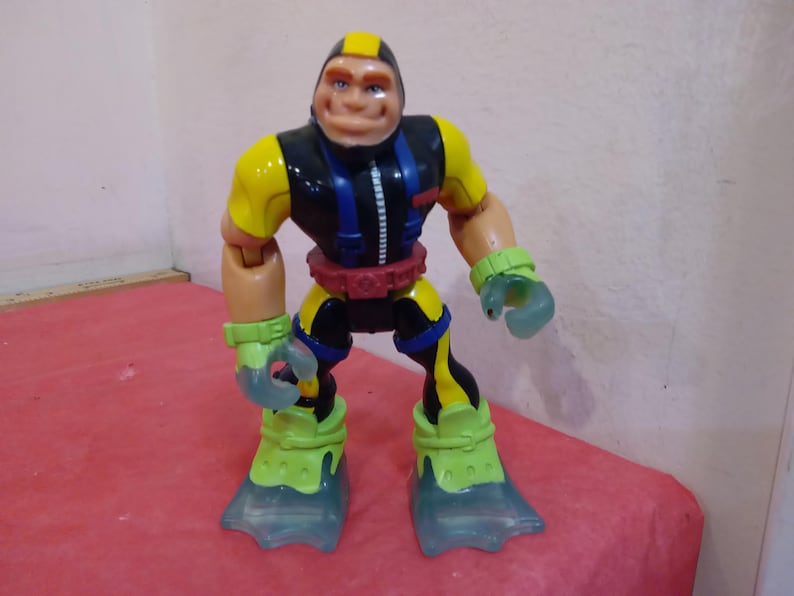 May include: A yellow, black, and blue action figure with a large head and flippers for feet. The figure is wearing a black wetsuit with a zipper and a yellow belt.