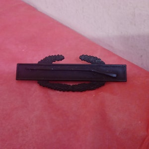 Puede incluir: Una insignia militar de metal negro con una corona de laurel y un rifle. La insignia es un símbolo del ejército español.