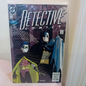 Può includere: Copertina del fumetto Detective Comics #647 con Batman e Robin. L'artwork ha uno sfondo viola e grigio, con il titolo "Detective Comics" in grandi lettere bianche. Il fumetto è in una custodia protettiva.