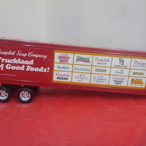 May include: A red toy truck trailer with the text "Campbell Soup Company Truckload of Good Foods!" and logos for various Campbell's products. The trailer has three sets of wheels.