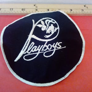 Peut inclure: Un écusson noir avec du texte brodé blanc et un logo. Le mot "Playboys" est écrit en script cursive. Le logo représente un lapin et un design stylisé. L'écusson a une bordure blanche.