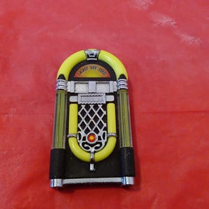 May include: A miniature jukebox with a yellow arch and a black base. The jukebox has silver accents and a diamond-shaped grill. The text "LIGHT my FIRE" is displayed on the top arch. A red jewel is in the center of the jukebox.