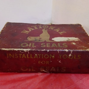 Op de afbeelding: Een vintage rode kartonnen doos met gouden letters die "National Oil Seals Installation Tools for Oil Seals" luidt. De doos heeft een afbeelding van een zeehond met het woord "National" erboven.