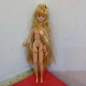 May include: A small doll with long, blonde hair and blue eyes. The doll is nude and standing on a red surface. A ruler is partially visible in the image. The doll is a vintage toy.