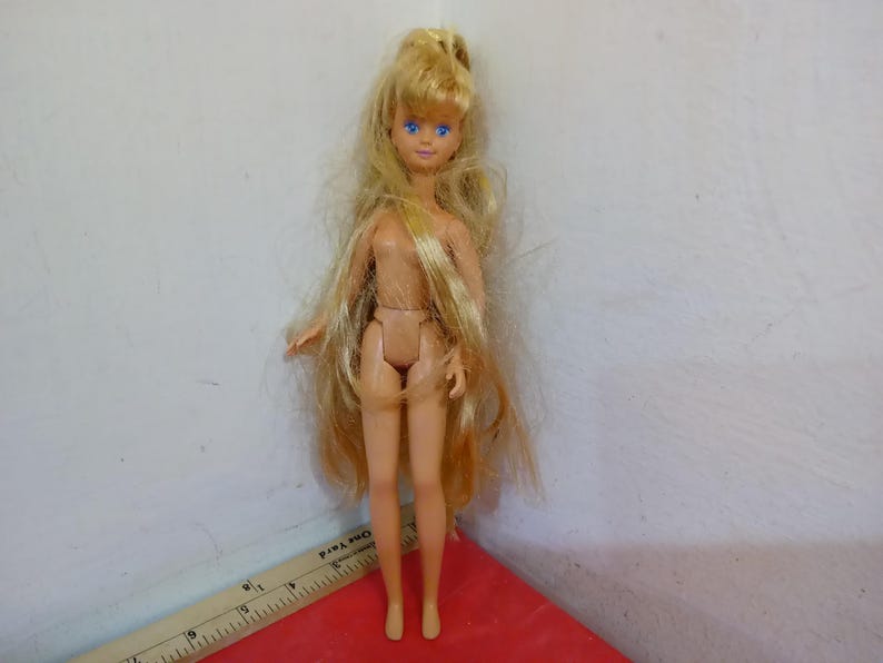 May include: A small doll with long, blonde hair and blue eyes. The doll is nude and standing on a red surface. A ruler is partially visible in the image. The doll is a vintage toy.
