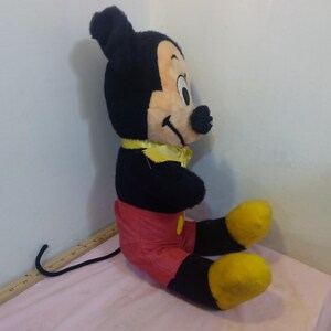 May include: A vintage Mickey Mouse plush toy, featuring black fur, a cream-colored face, and red shorts. The plush has yellow shoes and a yellow bow tie. The toy is sitting on a pink surface.