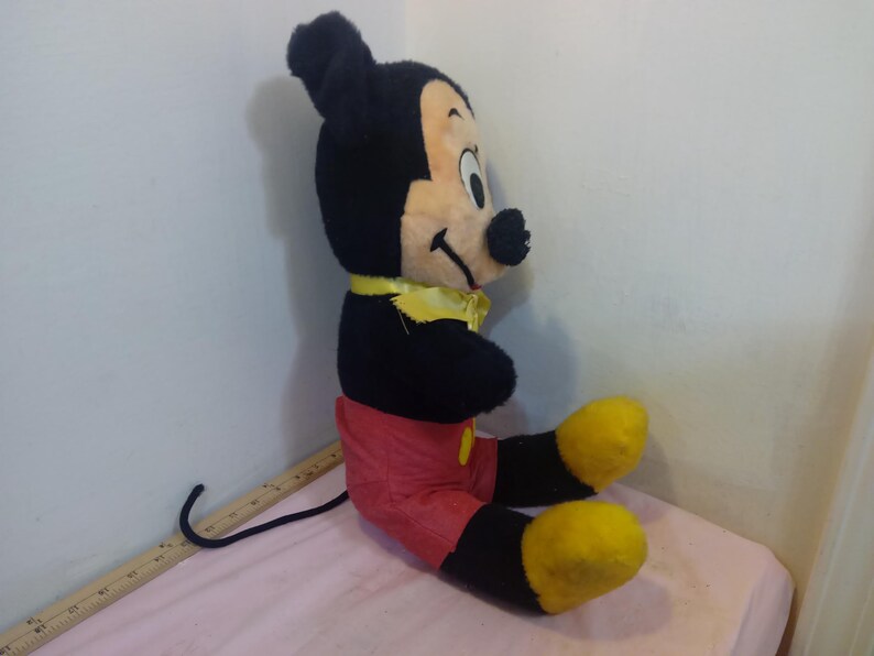 May include: A vintage Mickey Mouse plush toy, featuring black fur, a cream-colored face, and red shorts. The plush has yellow shoes and a yellow bow tie. The toy is sitting on a pink surface.