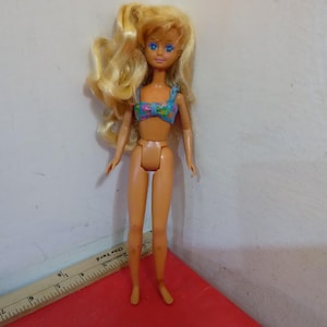 May include: A vintage doll with blonde hair and blue eyes, wearing a floral print bikini top. The doll is standing on a red surface. A wooden ruler with inch measurements is in the foreground.