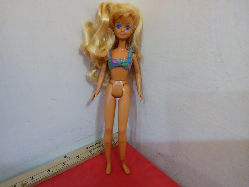 May include: A vintage doll with blonde hair and blue eyes, wearing a floral print bikini top. The doll is standing on a red surface. A wooden ruler with inch measurements is in the foreground.