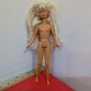 May include: A vintage doll with long blonde hair, blue eyes, and a small floral headband. The doll is nude, with articulated joints. It stands on a red surface, with a ruler visible in the foreground. The doll's skin tone is light.