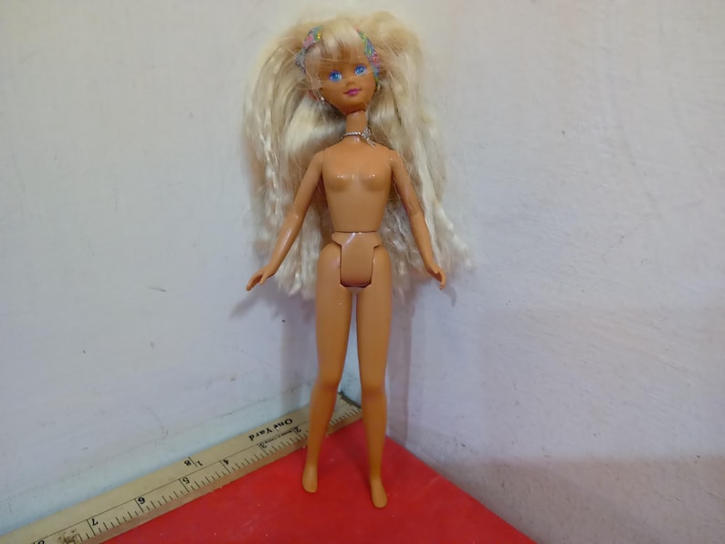 May include: A vintage doll with long blonde hair, blue eyes, and a small floral headband. The doll is nude, with articulated joints. It stands on a red surface, with a ruler visible in the foreground. The doll's skin tone is light.