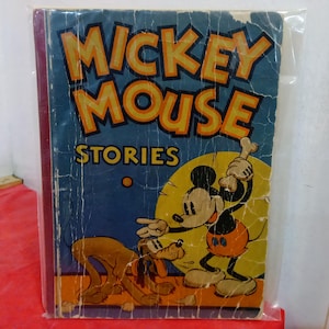 May include: Vintage "Mickey Mouse Stories" book. The cover features Mickey Mouse and Pluto with the title in yellow and orange against a blue background. The book is encased in clear plastic.