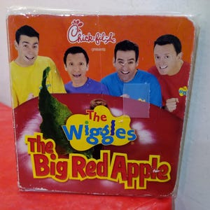 May include: A CD case for "The Wiggles: The Big Red Apple" featuring four smiling men in colorful shirts. The Chick-fil-A logo is at the top. The title is in yellow and red, with a green apple graphic.