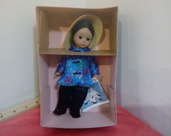 Könnte beinhalten: Eine kleine Puppe, die eine Jacke mit blauem und goldenem Muster, eine schwarze Hose und einen Strohhut trägt. Die Puppe befindet sich in einer Schachtel aus Pappe mit rosa Innenausstattung.